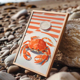 A framed image of a vibrant orange crab is displayed on a rocky beach, with a white plate featuring a small white object on top of it.