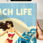 a hand holding a canvas print with a beach scene and a woman in a red swimsuit.