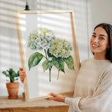 A woman is holding up a framed painting of a white hydrangea flower on a wooden table.