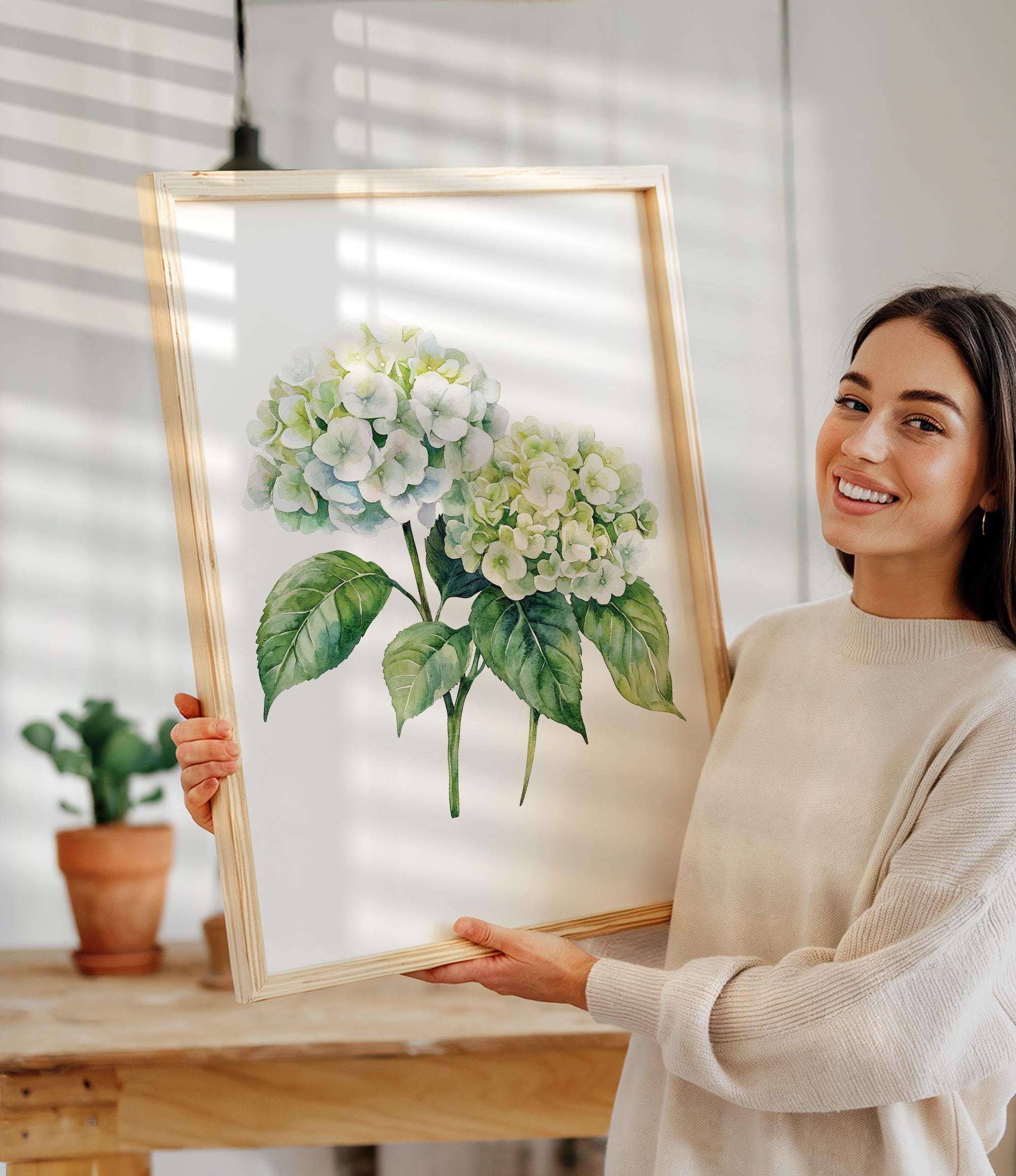 A woman is holding up a framed painting of a white hydrangea flower on a wooden table.