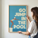 A woman is hanging a framed poster with the text "GO JUMP IN THE POOL" on a wall.