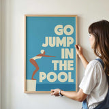 A woman is hanging a framed poster with the text "GO JUMP IN THE POOL" on a wall.