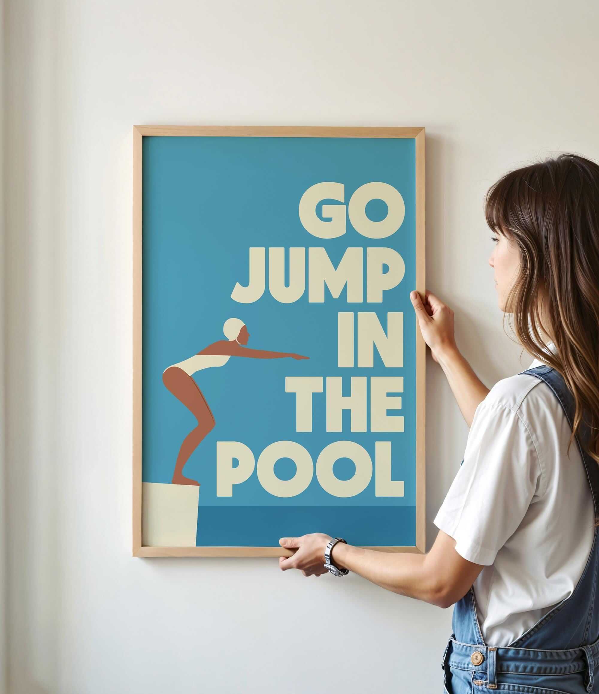 A woman is hanging a framed poster with the text "GO JUMP IN THE POOL" on a wall.