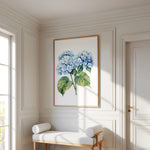 a white room with a large painting of blue hydrangeas hanging on the wall. In front of the painting, there is a white bench with two white cushions. The room has white walls and a wooden floor, creating a clean and elegant atmosphere.