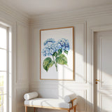 a white room with a large painting of blue hydrangeas hanging on the wall. In front of the painting, there is a white bench with two white cushions. The room has white walls and a wooden floor, creating a clean and elegant atmosphere.