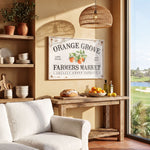 a cozy living room with a rustic wooden table, a white sofa, and a hanging woven light fixture. The table is adorned with various items, including a bowl of fruit, a loaf of bread, and a bottle of olive oil. Above the table, there is a framed sign that reads "Orange Grove Farmers Market Locally Grown Produce".