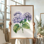 A person is holding up a framed painting of purple hydrangea flowers in a room with various potted plants and art supplies.