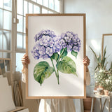 A person is holding up a framed painting of purple hydrangea flowers in a room with various potted plants and art supplies.