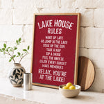 A framed sign with a red background and white text, placed on a white shelf next to a plant and a bowl of fruit.