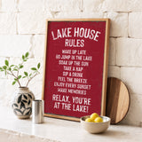 A framed sign with a red background and white text, placed on a white shelf next to a plant and a bowl of fruit.