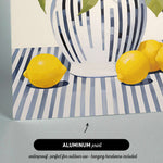 The image is an aluminum print depicting a still life arrangement of a vase with lemons and leaves on a striped tablecloth.