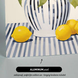 The image is an aluminum print depicting a still life arrangement of a vase with lemons and leaves on a striped tablecloth.