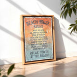 A framed sign with the text "BEACH HOUSE RULES" is displayed on a wooden floor, with a plant in the background.