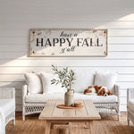 a cozy living room with a white sofa, a wooden coffee table, and a dog resting on the sofa. Above the sofa, there is a rustic wooden sign that reads "Have a HAPPY FALL y'all". The room has a white color scheme, with white walls and a white wicker chair. A vase with greenery is placed on the coffee table, and a rug covers the floor.