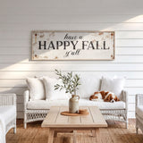 a cozy living room with a white sofa, a wooden coffee table, and a dog resting on the sofa. Above the sofa, there is a rustic wooden sign that reads "Have a HAPPY FALL y'all". The room has a white color scheme, with white walls and a white wicker chair. A vase with greenery is placed on the coffee table, and a rug covers the floor.