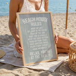 A woman is sitting on a beach towel, holding a sign that reads "BEACH HOUSE RULES" in front of her.