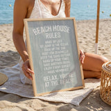 A woman is sitting on a beach towel, holding a sign that reads "BEACH HOUSE RULES" in front of her.