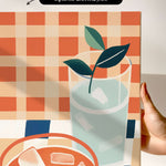 The image is a hand holding a canvas print featuring a glass of water with ice cubes, a plant, and a bowl of food.