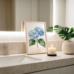 a framed painting of blue hydrangeas on a bathroom counter, with a lit candle in a white vase and a potted plant nearby.
