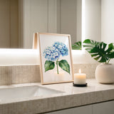 a framed painting of blue hydrangeas on a bathroom counter, with a lit candle in a white vase and a potted plant nearby.