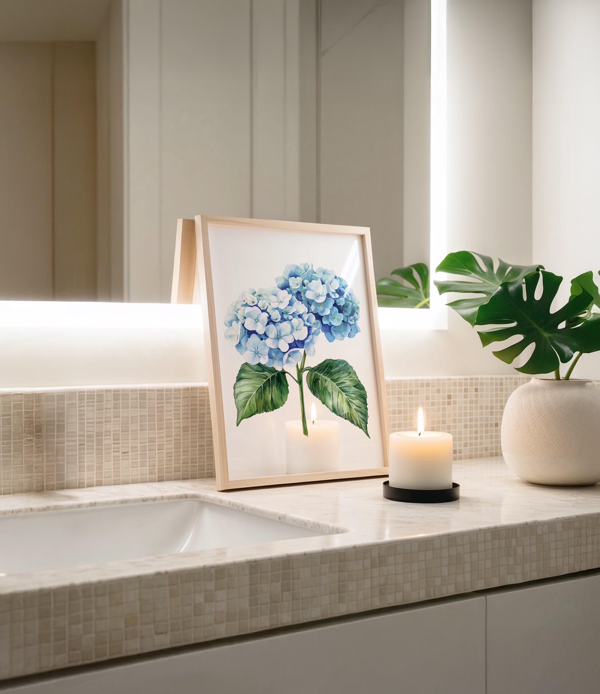 a framed painting of blue hydrangeas on a bathroom counter, with a lit candle in a white vase and a potted plant nearby.