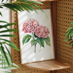 A framed watercolor painting of pink hydrangeas is displayed on a wooden shelf, surrounded by lush green plants.