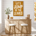 a cozy and inviting bar area with a wooden bar counter, two white stools, and a framed sign that reads "Let's JUMP in the LEAVES". The bar counter is adorned with a vase of greenery and a basket, adding a touch of nature to the space. The overall atmosphere of the image is warm and welcoming, inviting patrons to enjoy their time at the bar.