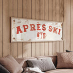a wooden wall with a framed sign that reads "APRÈS SKI", and there is a couch with pillows in the foreground.