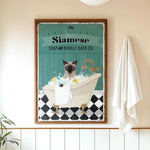 A framed vintage-style poster of a Siamese cat sitting in a bathtub, surrounded by bubbles, with a towel hanging on the wall next to it.