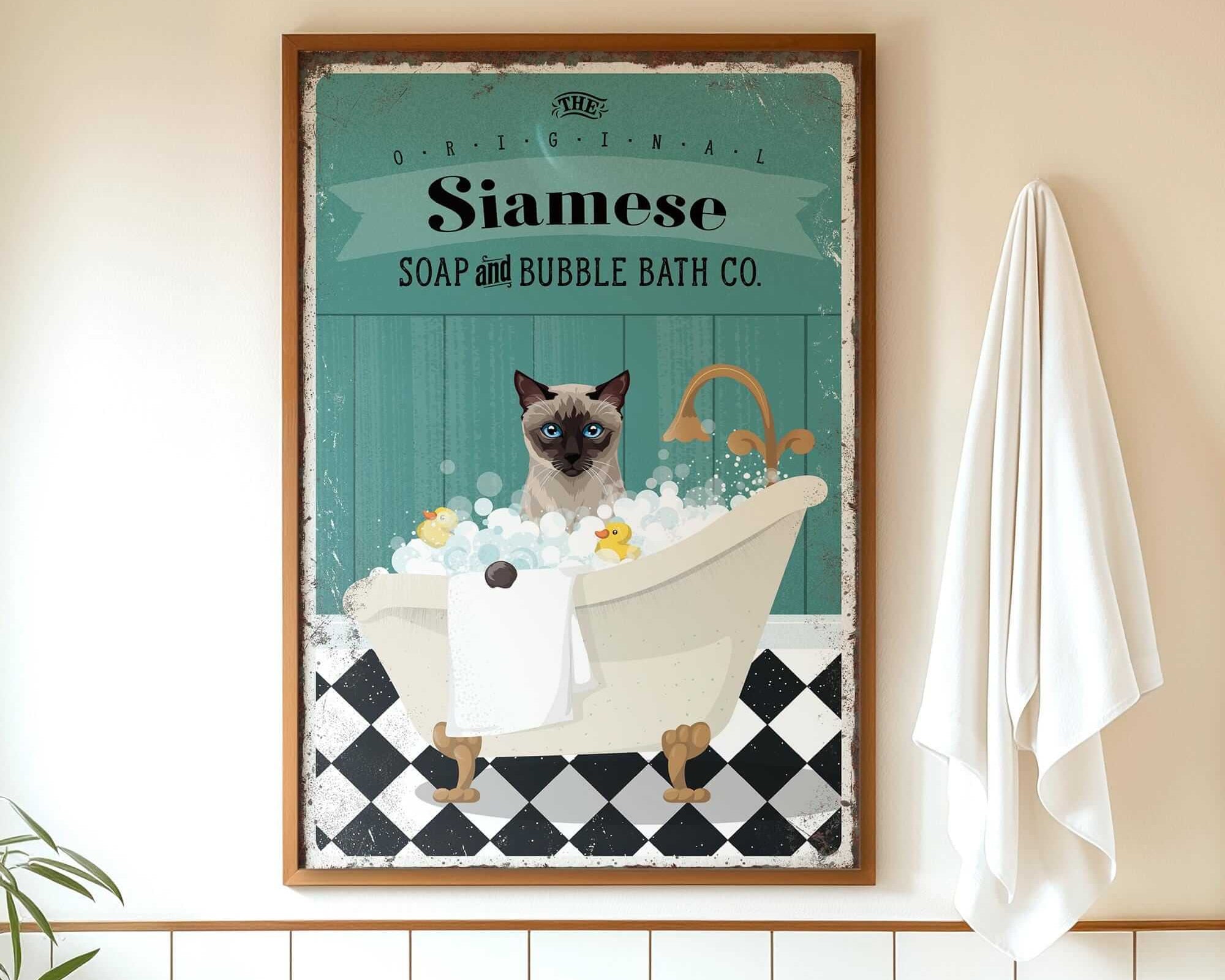 A framed vintage-style poster of a Siamese cat sitting in a bathtub, surrounded by bubbles, with a towel hanging on the wall next to it.