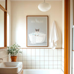 A bathroom with a framed poster of a cat in a bathtub, a towel rack, and a potted plant.