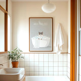 A bathroom with a framed poster of a cat in a bathtub, a towel rack, and a potted plant.