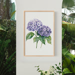 a framed watercolor painting of purple hydrangeas hanging on a wall, surrounded by lush greenery and tropical plants.