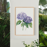 a framed watercolor painting of purple hydrangeas hanging on a wall, surrounded by lush greenery and tropical plants.