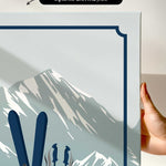 a hand holding a framed print of a mountain landscape with skis and ski poles.