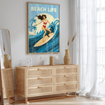 a framed poster of a woman surfing on a wave, with the text "BEACH LIFE" written above it. The poster is displayed on a wall above a wooden dresser with drawers.
