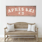 a rustic wooden bench with two striped pillows, set against a white wall with a framed sign above it that reads "APRES SKI".