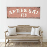 a rustic wooden bench with two striped pillows, set against a white wall with a framed sign above it that reads "APRES SKI".