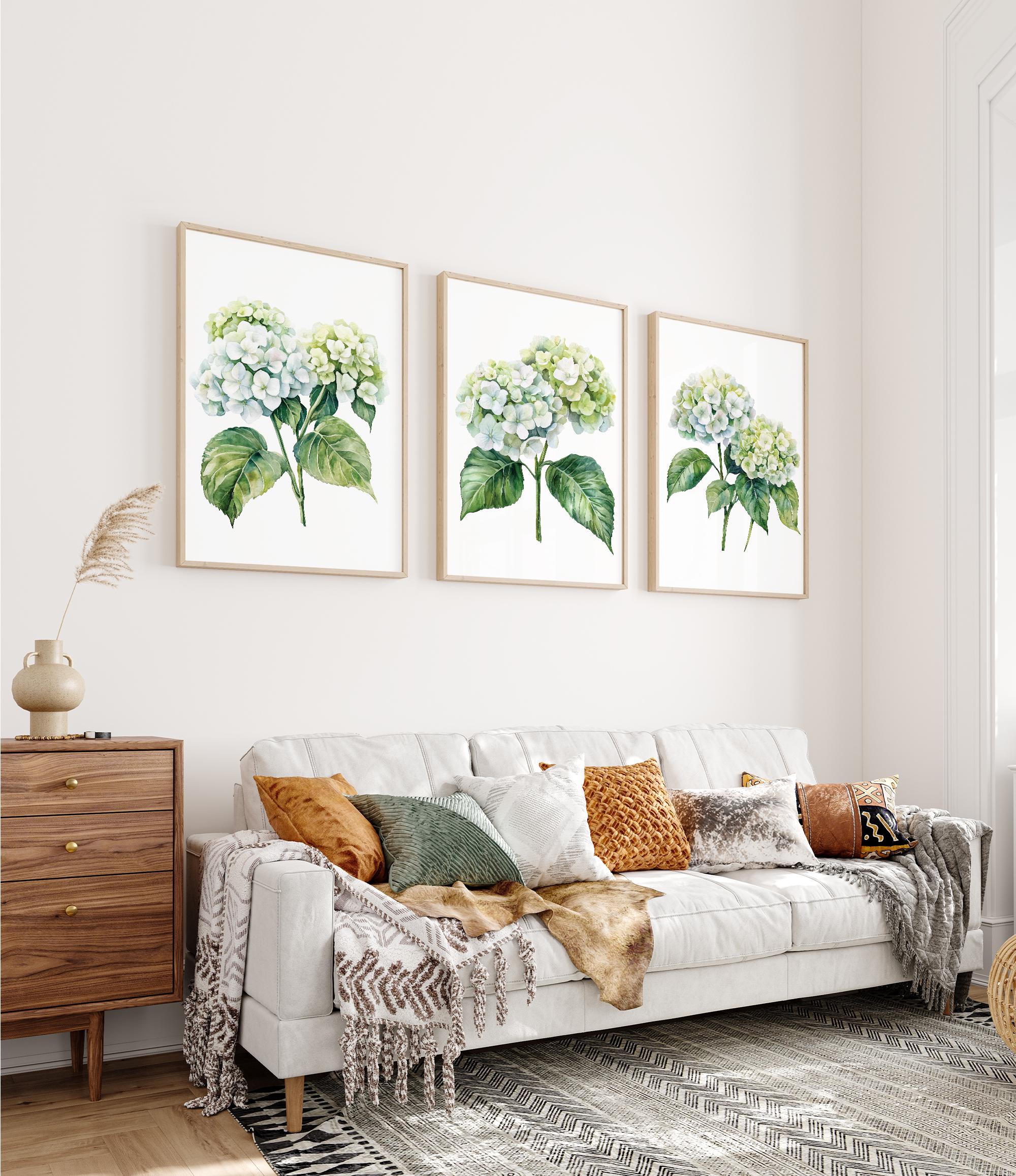 a cozy living room with a white sofa adorned with throw pillows and a blanket. Above the sofa, there are three framed watercolor paintings of hydrangeas hanging on the wall. The room also includes a wooden dresser and a rug, creating a warm and inviting atmosphere.