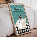 A framed poster depicting a Siamese cat sitting in a bathtub filled with bubbles, with a towel and a sink nearby.