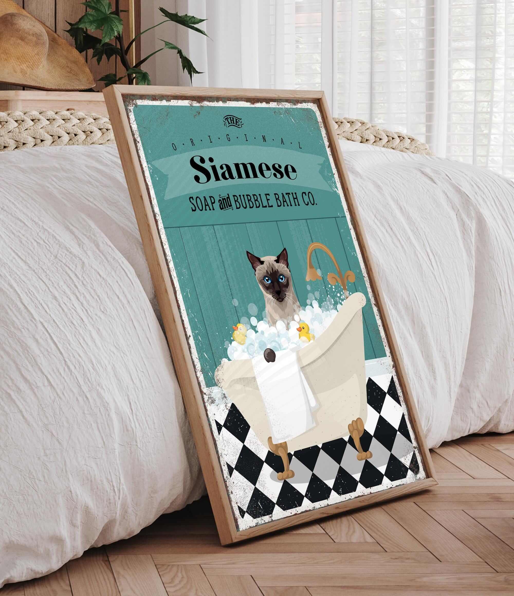 A framed poster depicting a Siamese cat sitting in a bathtub filled with bubbles, with a towel and a sink nearby.