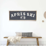 a wooden bench with a gray and white throw blanket on it, and a framed sign above it that says "APRES SKI".