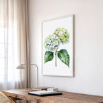 a wooden desk with a lamp, a framed watercolor painting of a green hydrangea flower, and a window with sheer curtains.