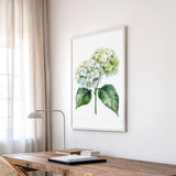 a wooden desk with a lamp, a framed watercolor painting of a green hydrangea flower, and a window with sheer curtains.