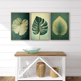 a white console table with three framed leaf artworks displayed above it. The leaves are green and yellow, creating a harmonious color palette. The table also has a glass bowl and a woven basket on it, adding a touch of natural elements to the interior design.