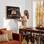 A woman hangs a framed sign on a white wall in a cozy living room, with a wooden dining table and chairs nearby.