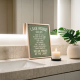 A framed sign on a bathroom counter displaying "Lake House Rules" and a lit candle, with a potted plant and a sink in the background.