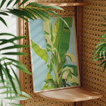 a framed painting of a banana tree on a wooden shelf, surrounded by lush green plants.