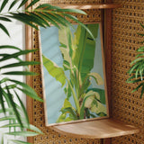a framed painting of a banana tree on a wooden shelf, surrounded by lush green plants.