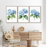 a living room with a wooden dresser, a woven basket, and a woven chair. Above the dresser, there are three framed watercolor paintings of blue hydrangeas hanging on the wall.